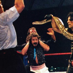 Alice Cooper spooks the referee with Damien the Snake at WrestleMania III while Jake Roberts applies a Full Nelson on Jimmy Hart.