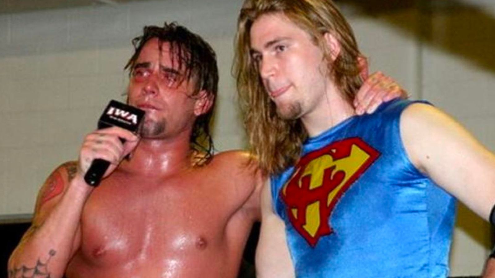 Chris Hero appears alongside CM Punk during an IWA promo, sporting his signature Superman-inspired "CH" insignia. Before adopting the name Hero, he wrestled under a controversial persona that sparked backlash. The Superman motif became a defining part of his rebrand, blending comic book influence with indie wrestling flair.