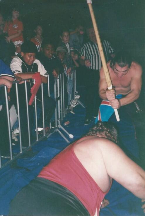Chris Michaels (Bill Pierce) moments before striking Typhoon (Fred Ottman) with a bamboo stick during a Tommy Dee show, showcasing the hard-hitting style that earned respect from wrestling’s giants. 