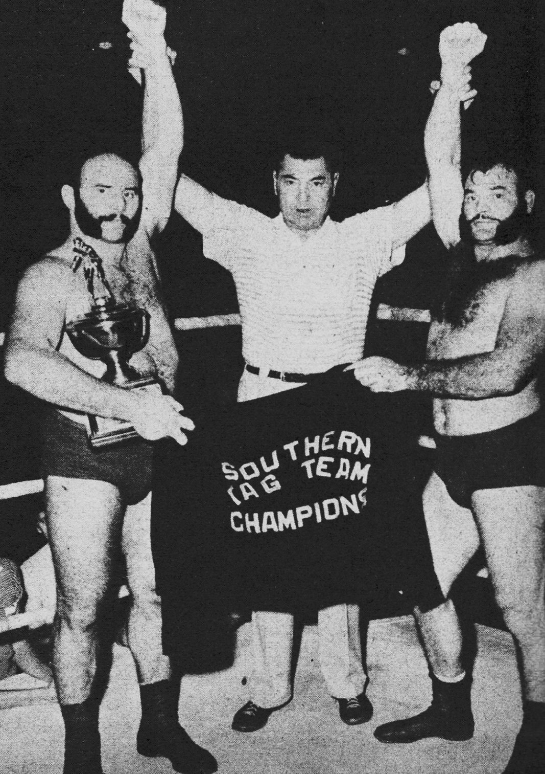 John and Al Smith, flanked by referee Jack Dempsey, posed with the Southern Tag Team trophy and their championship jackets after a title win in Nashville, Tennessee, on September 8, 1953, proof that Charlotte’s top heel brothers were not just drawing heat, they were taking home hardware. 